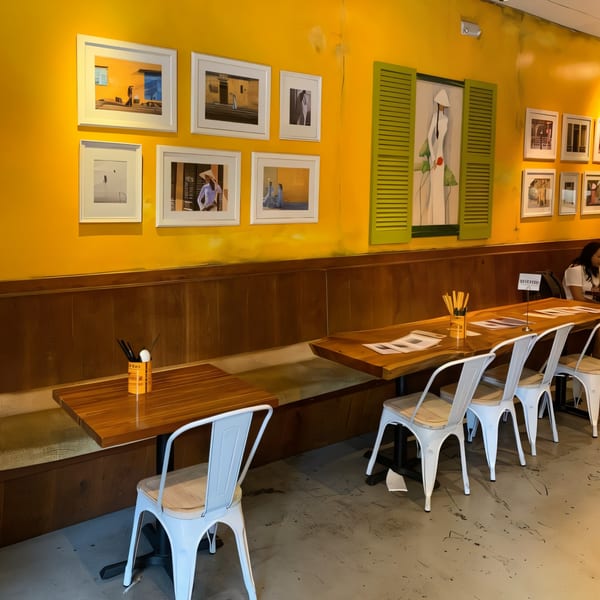 Interior of Anh's Kitchen with yellow walls, framed art, wooden tables, and white chairs.