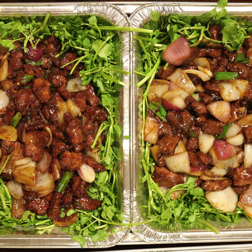 Two foil containers filled with dark glazed meat, onions, and green herbs.