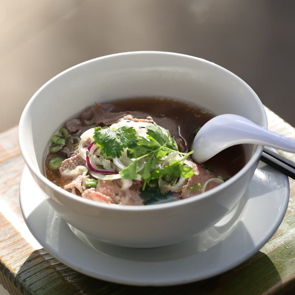 A bowl of Anh's Kitchen's Vietnamese pho with beef, onions, and cilantro, on a wooden table.