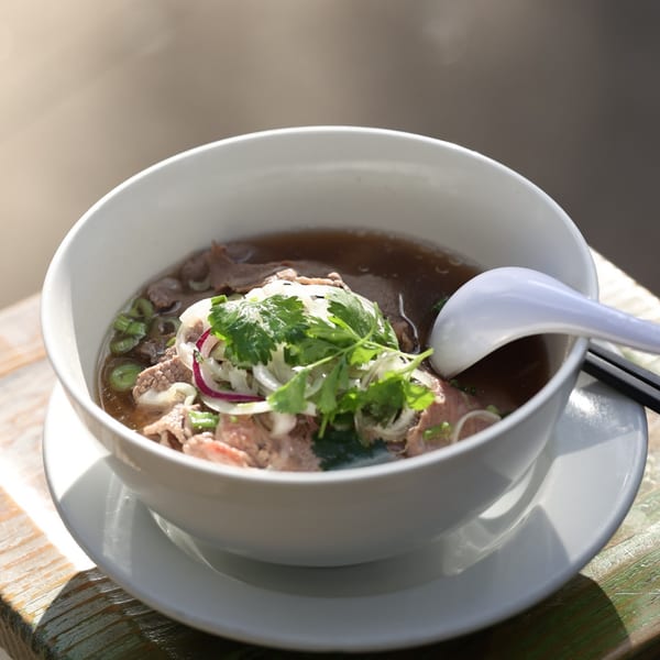 A bowl of Vietnamese pho with beef, onions, and cilantro, with a spoon and chopsticks.