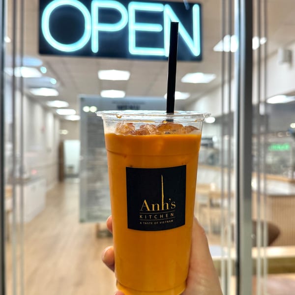 Hand holding an Anh's Kitchen iced Thai tea with a neon "OPEN" sign in the background.