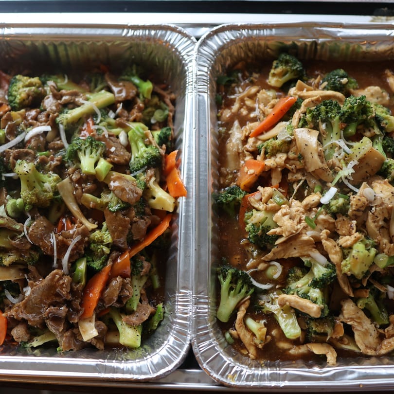 Two foil trays filled with stir-fried beef and chicken with broccoli, carrots, and mushrooms.