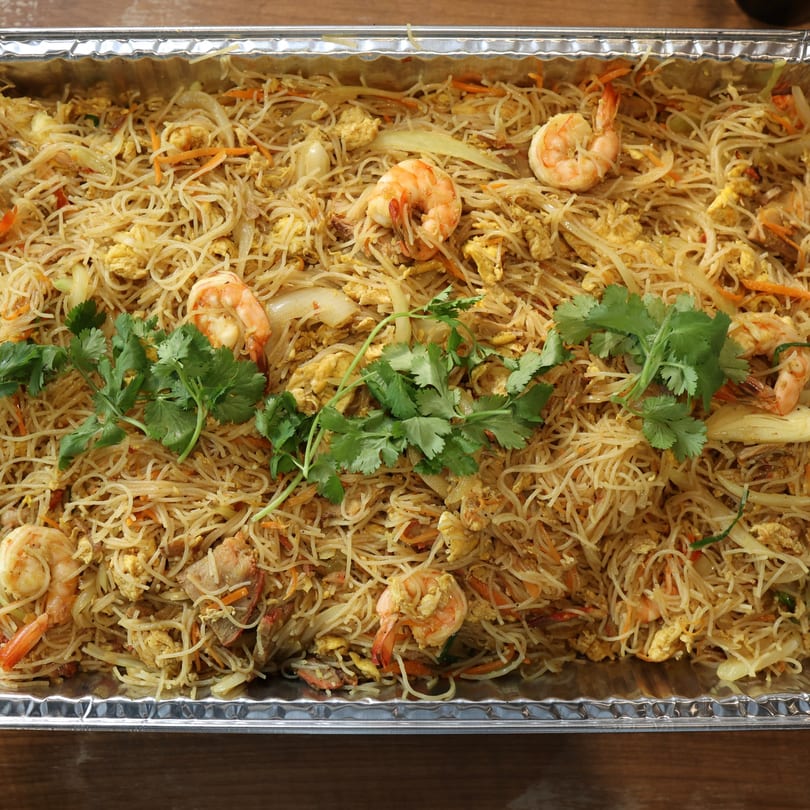 A large aluminum tray filled with Singapore noodles, shrimp, vegetables, and cilantro.