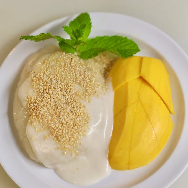 Mango sticky rice with coconut milk, sesame seeds, and a mint garnish on a white plate.