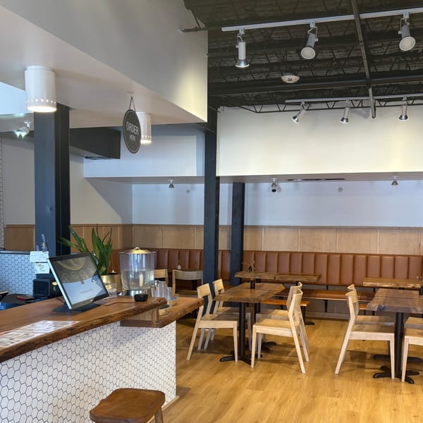 Modern restaurant interior with a wooden counter, white hexagonal tiles, light wood tables, and brown booth seating.