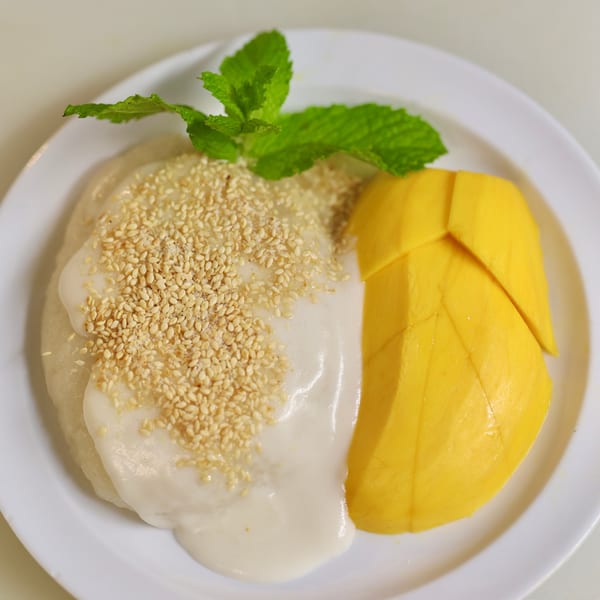 Mango sticky rice dessert with coconut milk, sesame seeds, and a mint garnish on a white plate.