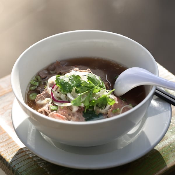 A bowl of Anh's Kitchen pho with beef, onions, and cilantro, served with a spoon and chopsticks.