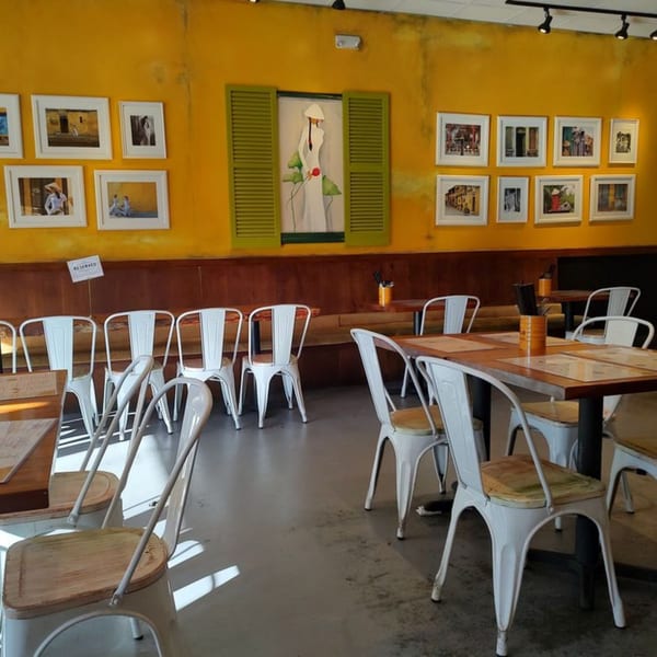 Anh's Kitchen dining room with yellow walls, Vietnamese art, and white metal chairs at wooden tables.