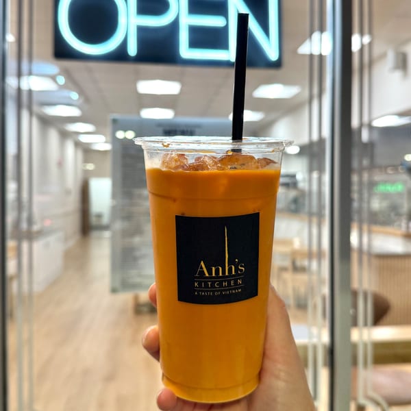 Hand holding an Anh's Kitchen iced Thai tea with a black straw, in front of an "OPEN" sign.
