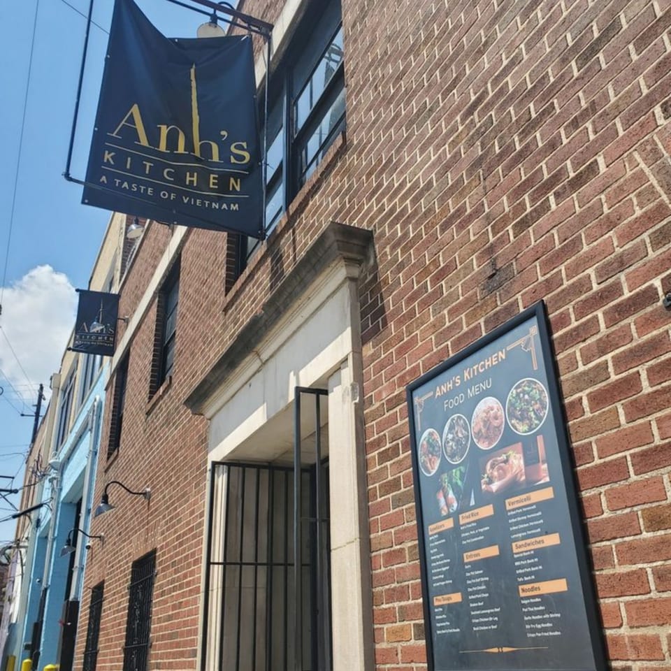 Anh's Kitchen sign and outdoor menu board on a brick building facade under a blue sky.