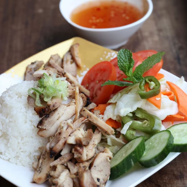 Grilled chicken with rice, sliced tomatoes, pickled vegetables, and cucumbers, with a side of sauce.