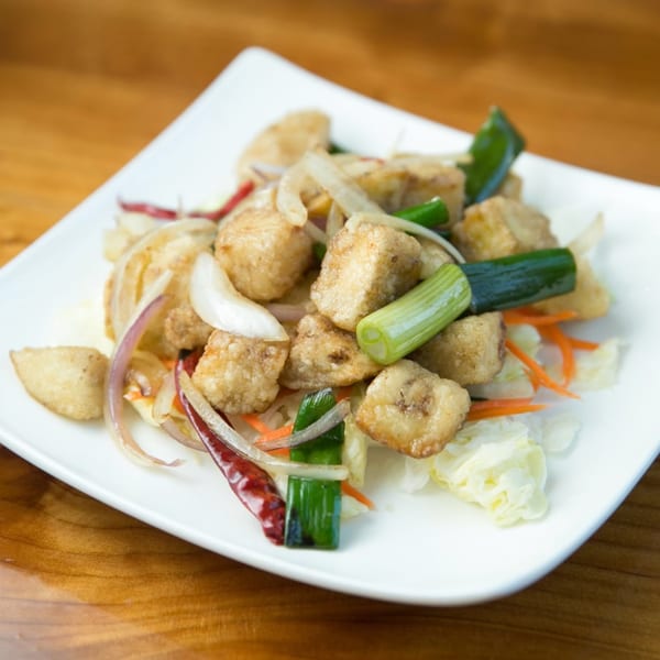 Crispy fried tofu with red onions, green onions, and red chili peppers on a white plate.